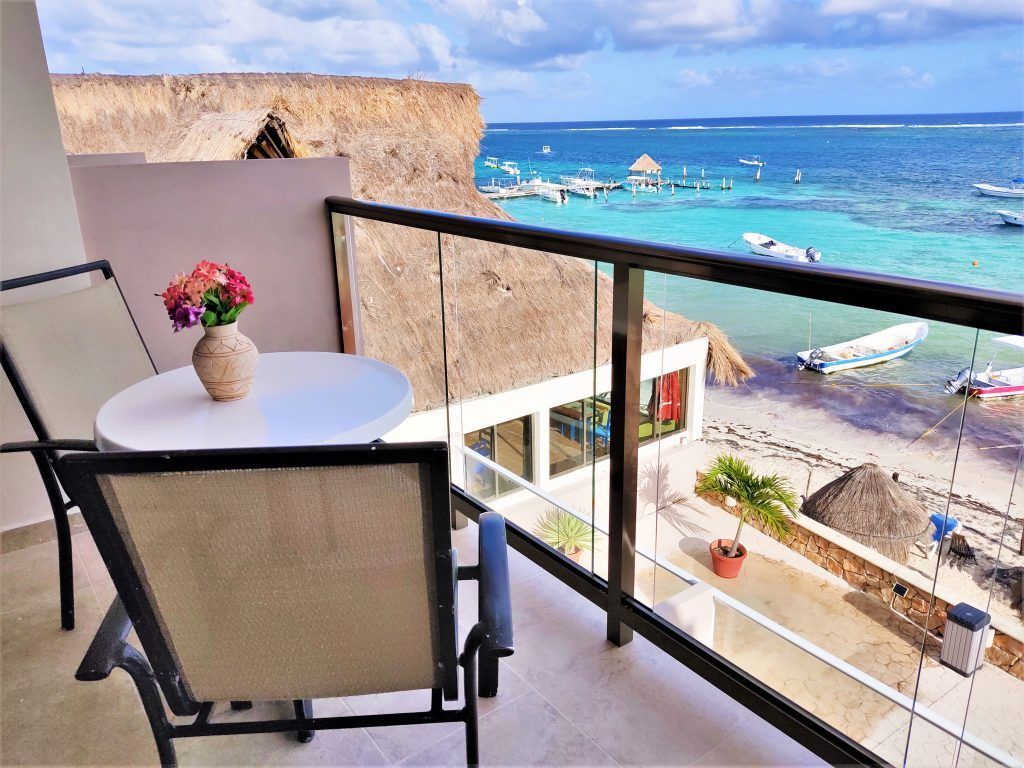 Balcony with table, chairs, vase of flowers, overlooking turquoise ocean and beach.