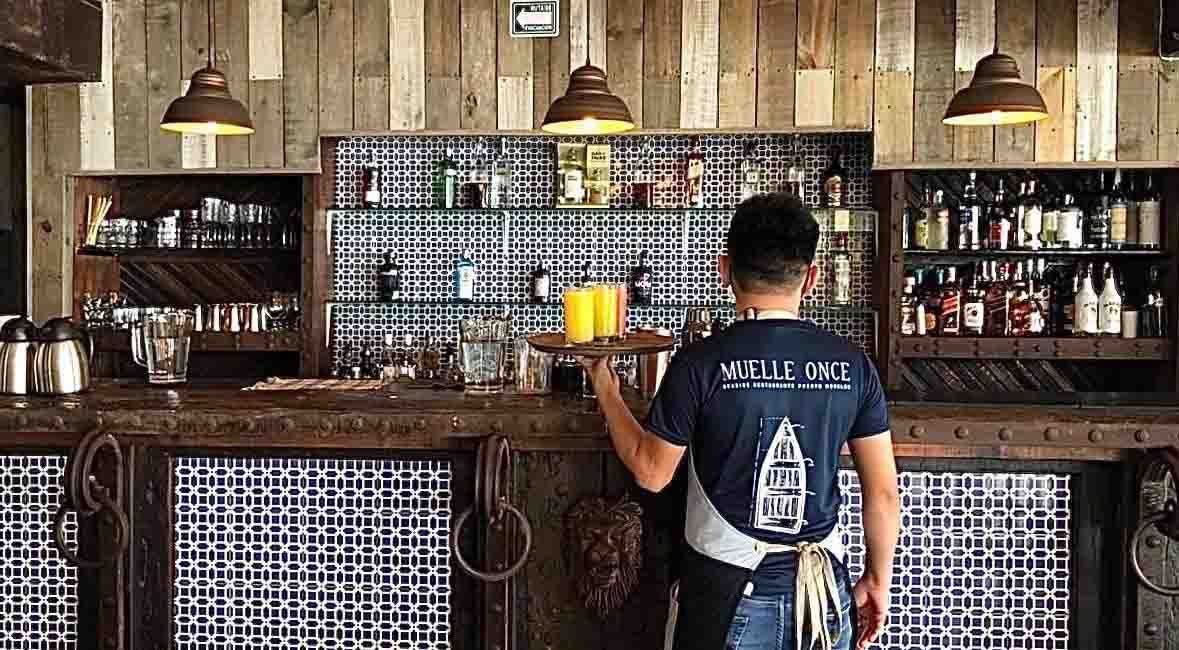 Bartender holding tray, standing behind a rustic bar with shelves of liquor bottles.