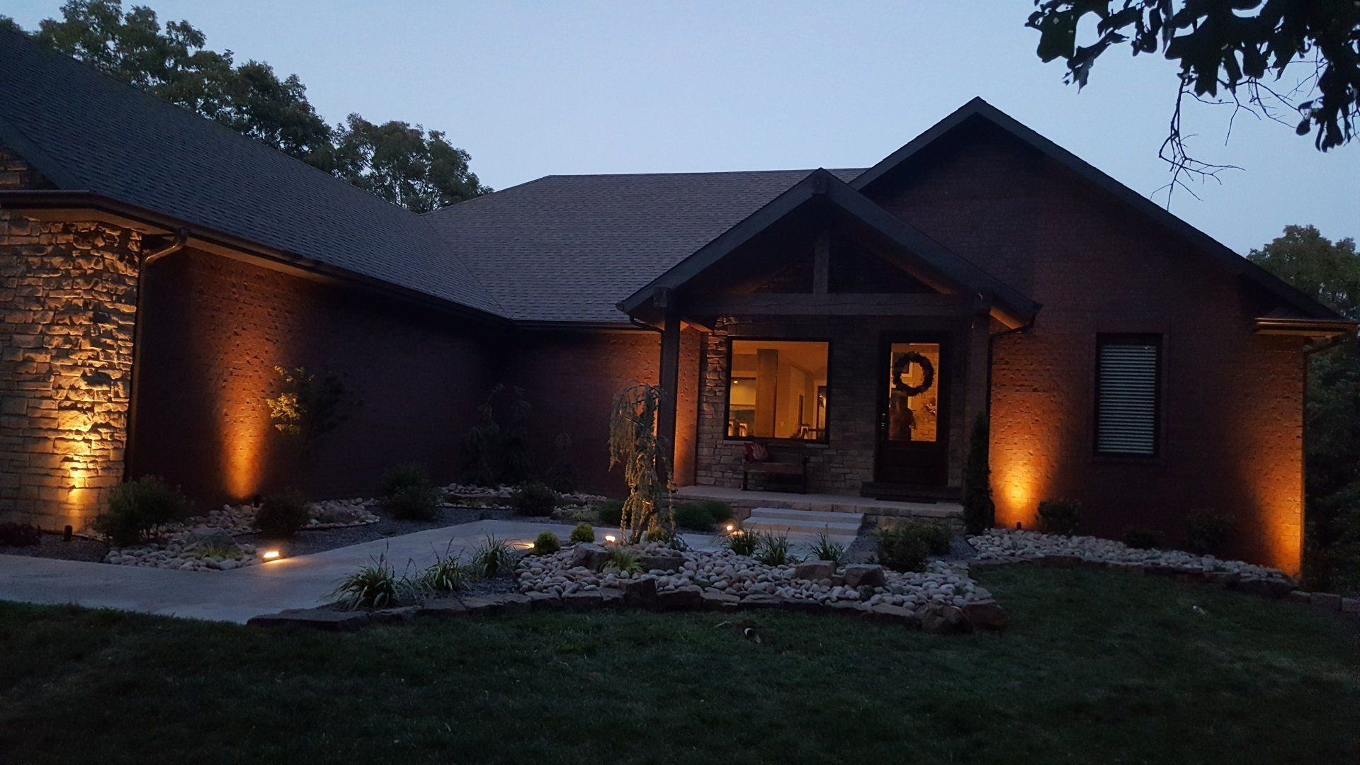 House exterior at dusk, illuminated by spotlights, with a stone facade and front porch.