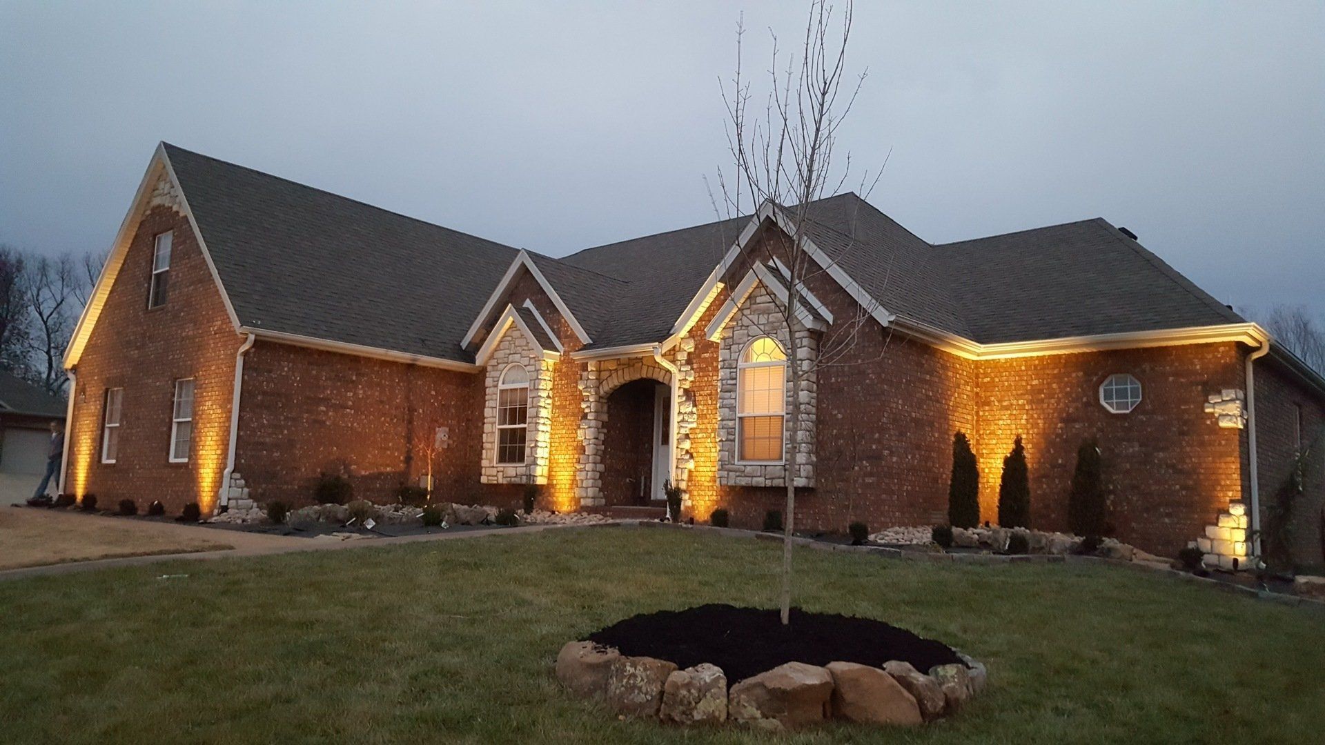 Brick house at dusk, illuminated by warm outdoor lights. Landscaping and grass in front.