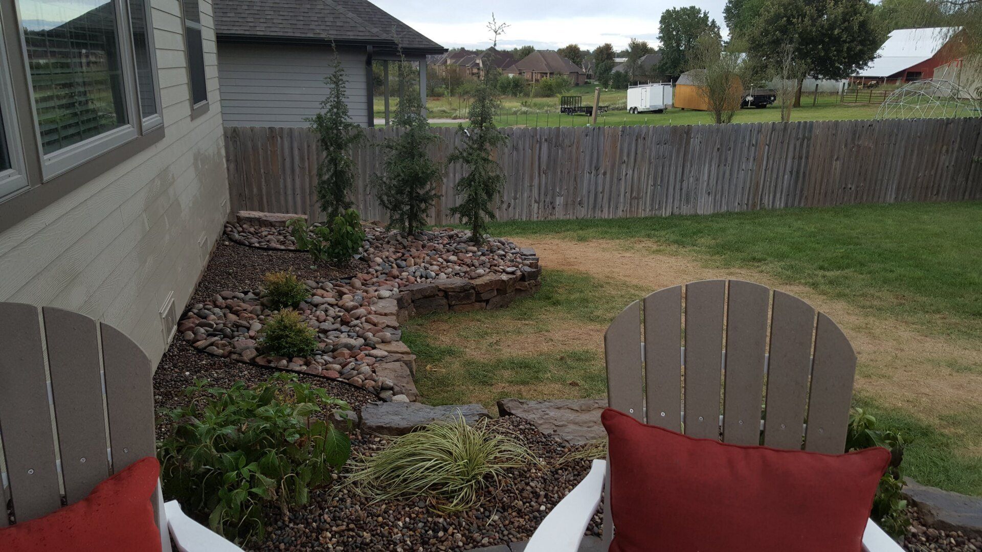 Two Adirondack chairs with red pillows on a patio, overlooking a garden and fence.