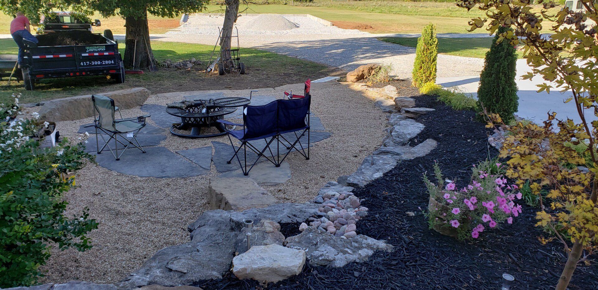 Backyard fire pit area with gravel, rocks, chairs, and landscaping.