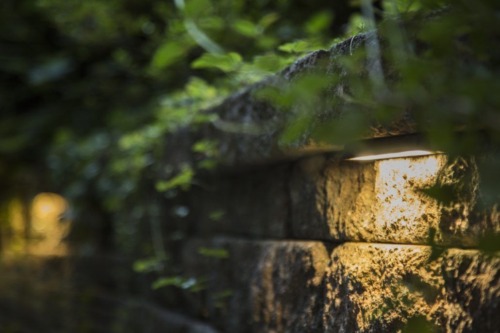 Stone wall with built-in lights, partially obscured by green foliage, casting a warm glow.