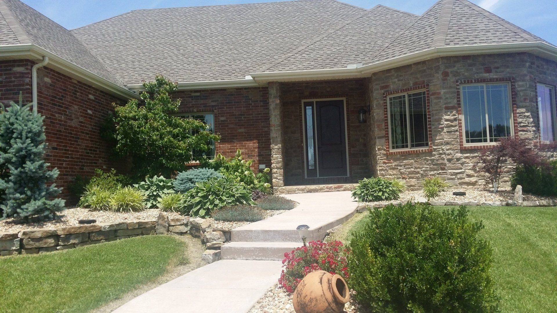 A brick and stone house with a curved pathway, lush landscaping, and a patterned roof.