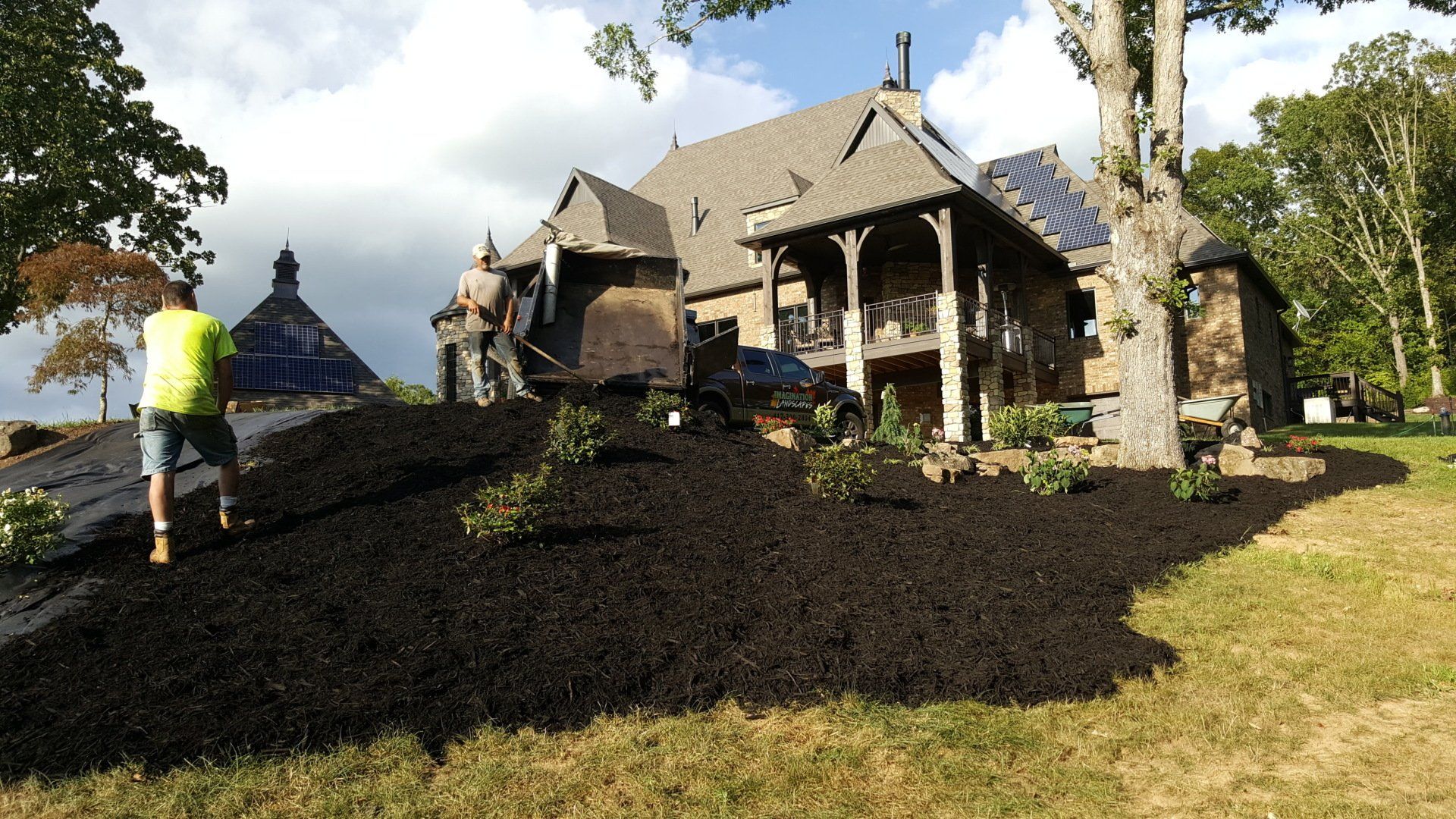Landscapers mulching a large house with a stone and wood facade on a sunny day.