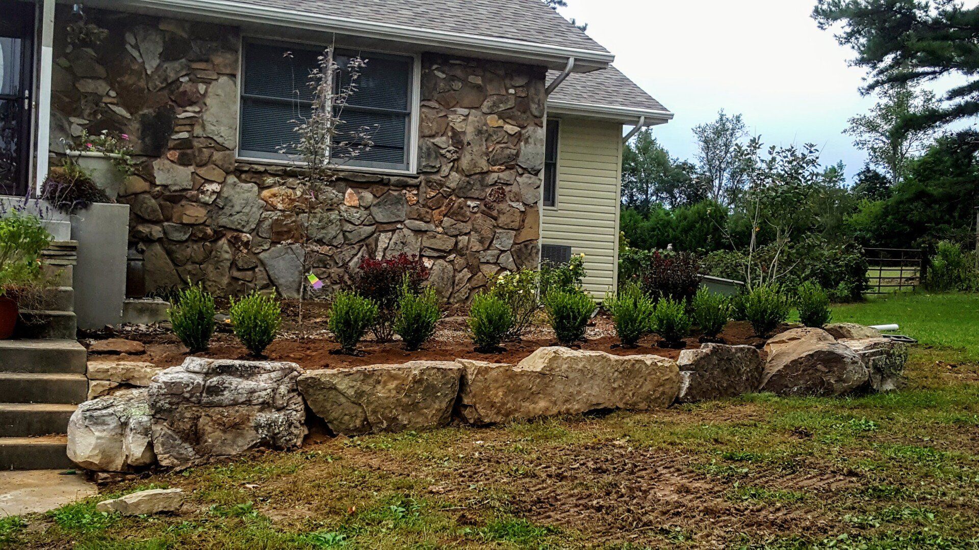 Stone house with a rock retaining wall, featuring trimmed bushes and a green lawn.