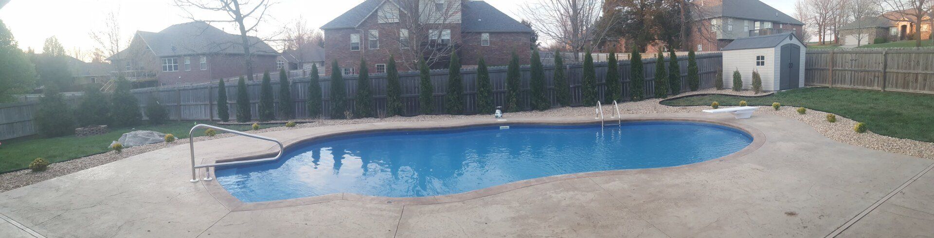A backyard with a blue swimming pool surrounded by concrete, green grass, and trees.