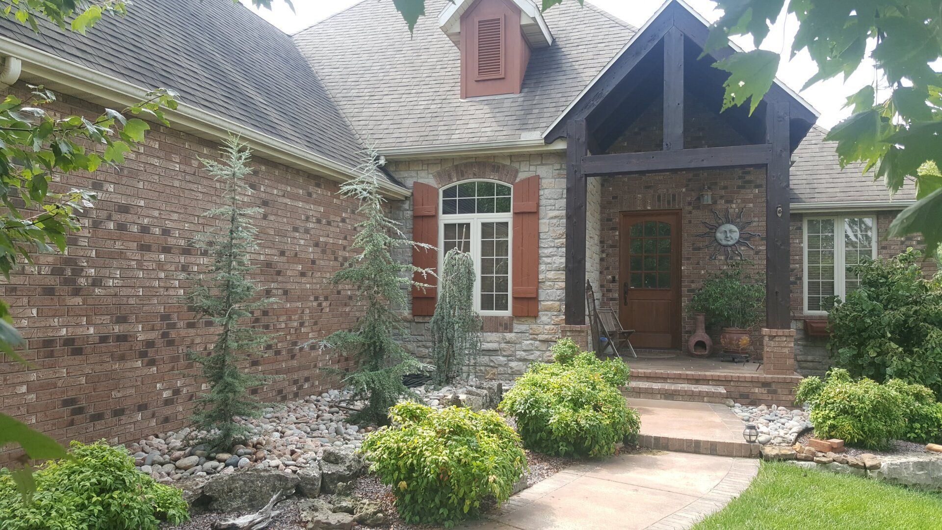 Stone and brick house with dark roof, shutters, and porch. Landscaping in front.