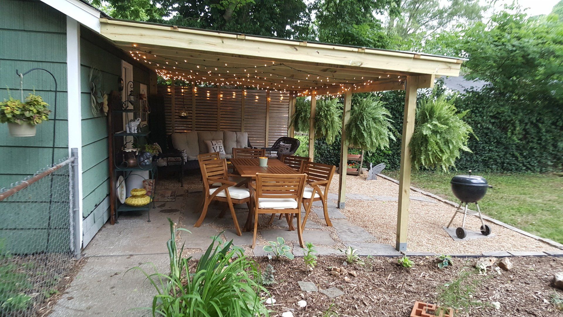 Outdoor dining area under a wooden awning, featuring a table, chairs, string lights, and a grill.