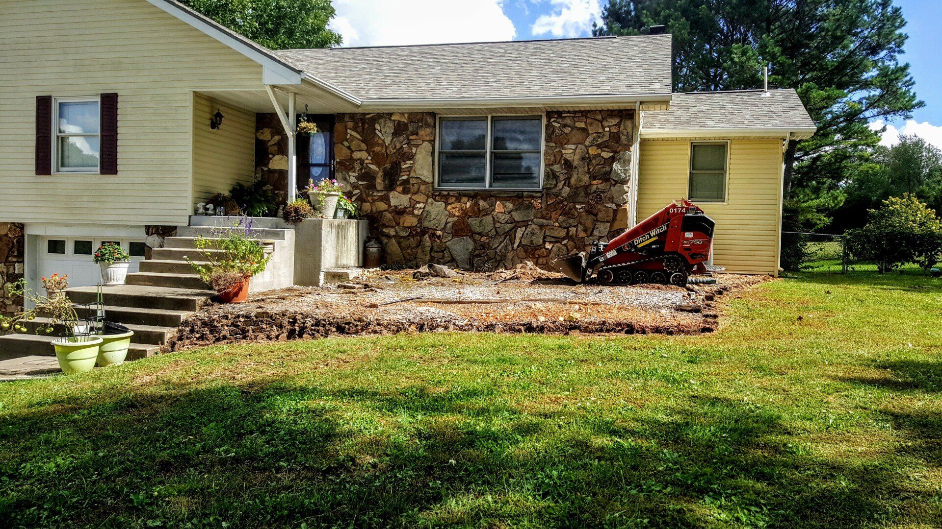 House exterior with foundation work in progress, rock facade, yellow siding, green lawn. A small excavator is present.
