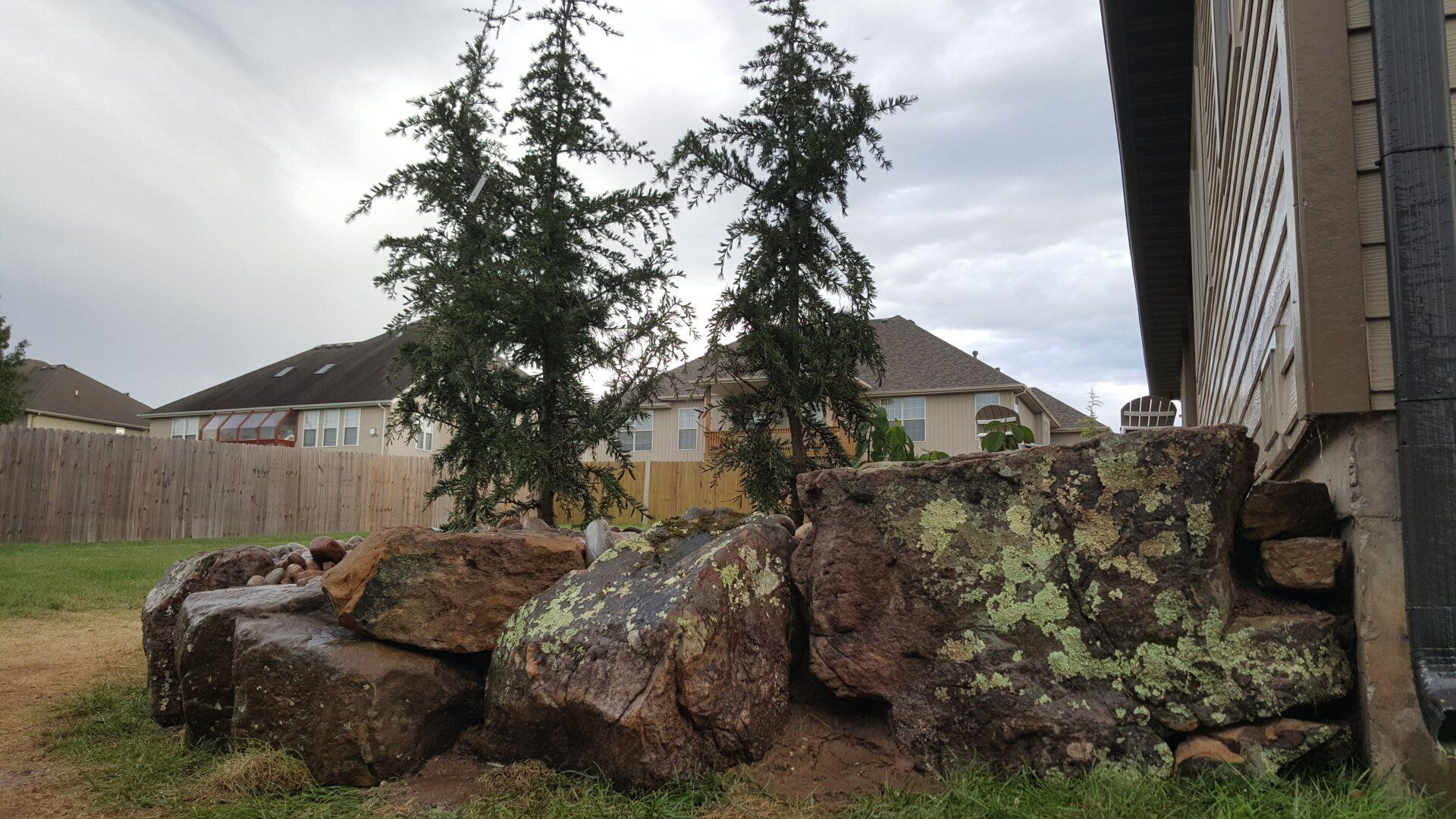 Stone retaining wall with two evergreen trees and a house in the background on a cloudy day.