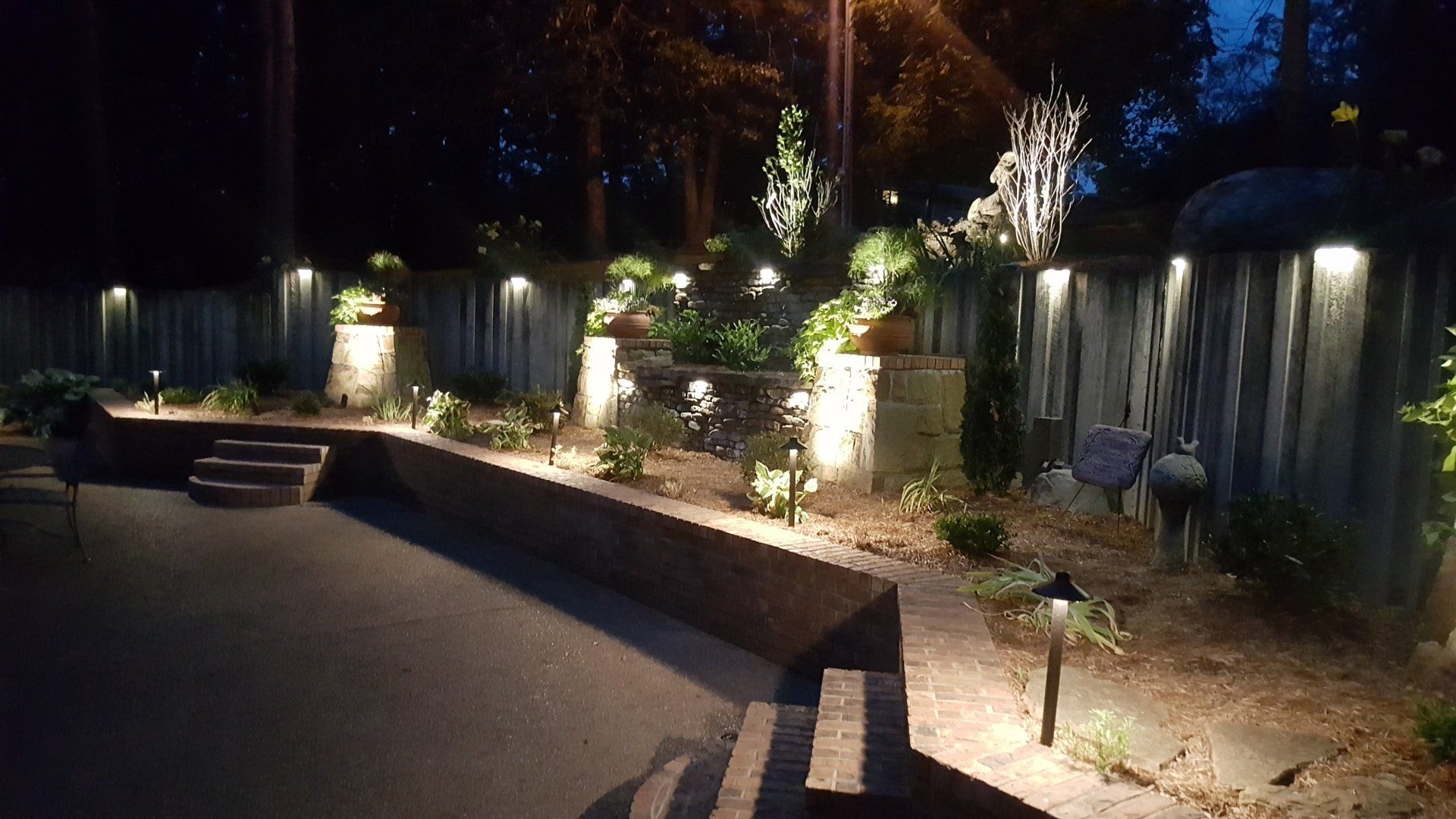 Nighttime view of a brick and wooden fence with outdoor lighting illuminating plants and steps.