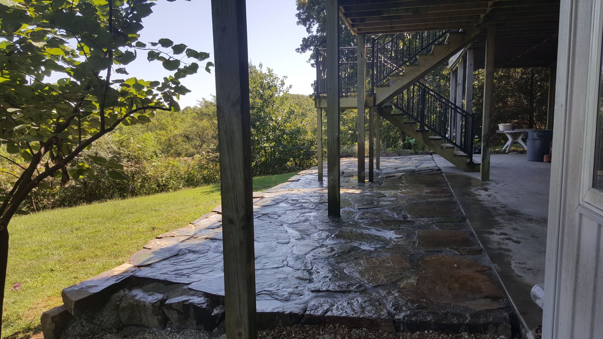 Wet concrete patio under a deck, with support beams. Lush greenery in the background.