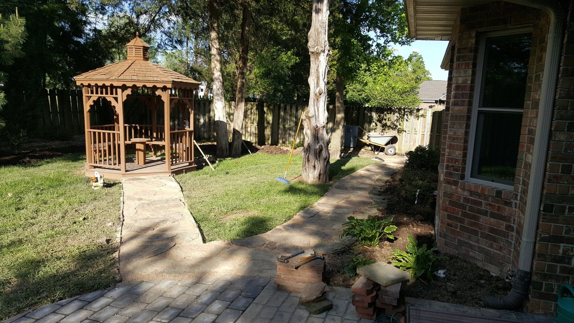 Gazebo in a backyard with a brick path, adjacent to a brick house. Green grass, trees, and wooden fence.