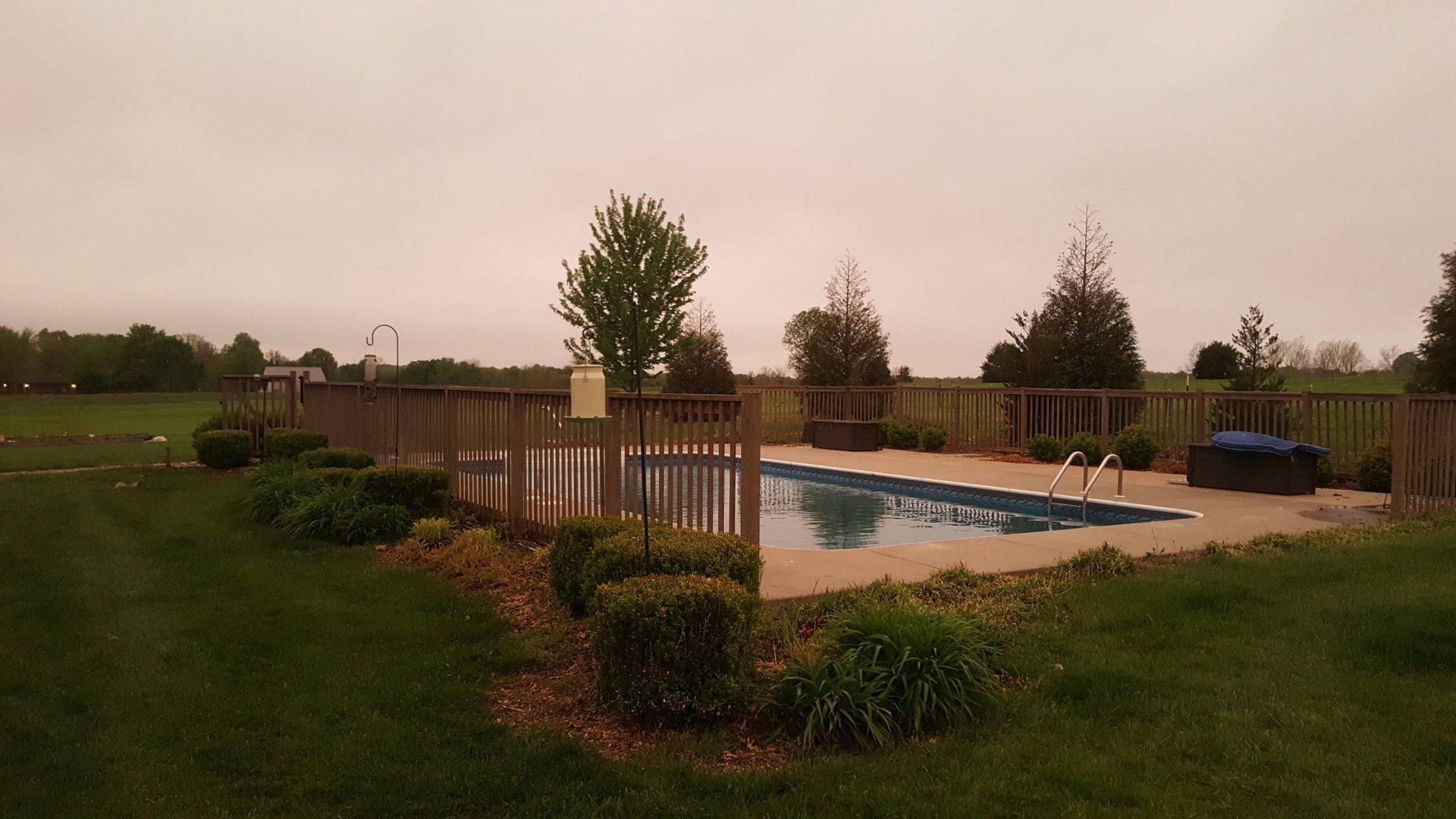 Pool surrounded by a wooden fence, bushes, and green grass under a hazy, brown sky.