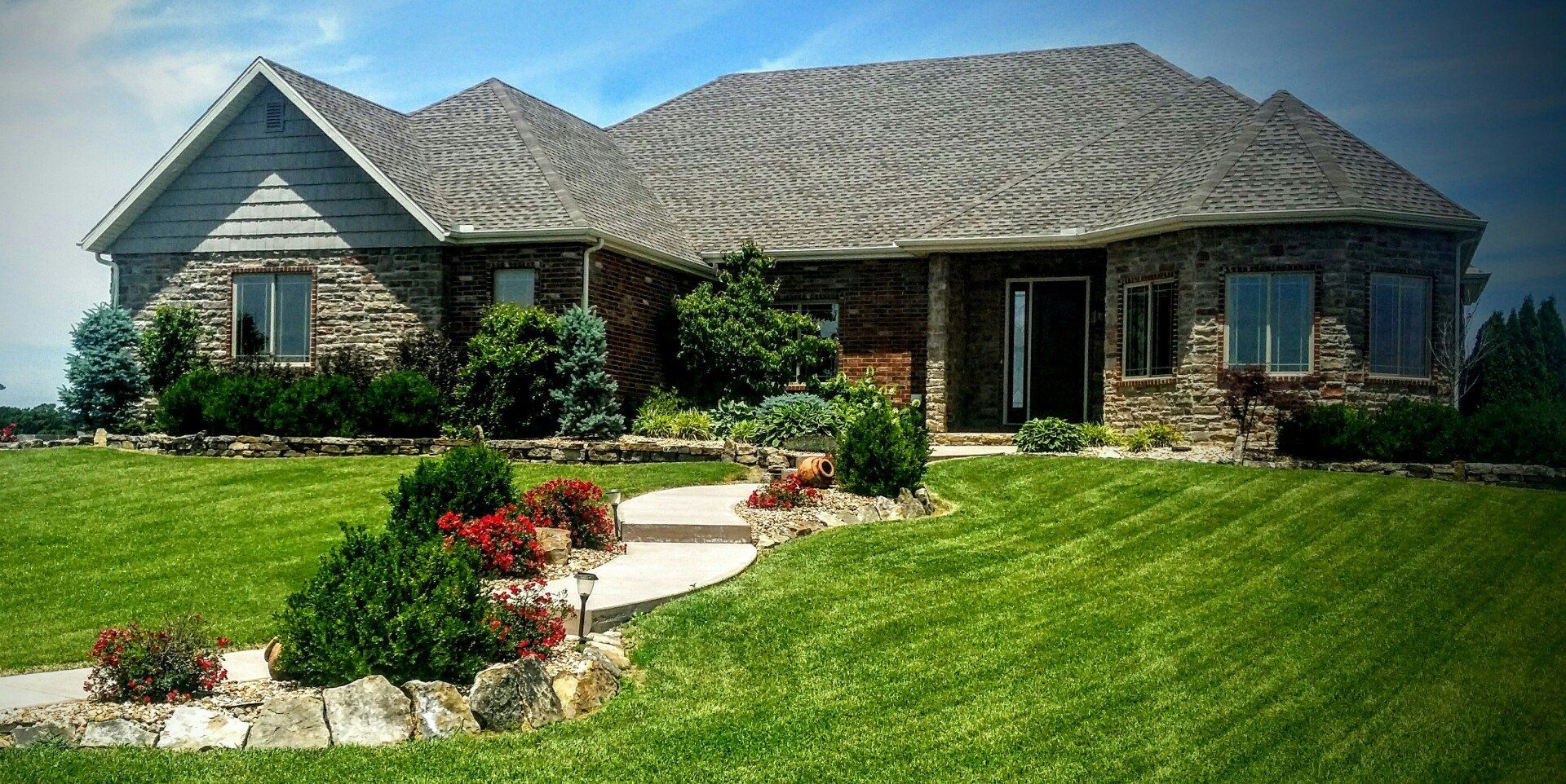 Stone-clad house with a wavy roof, lush green lawn, and landscaped pathway, under a blue sky.