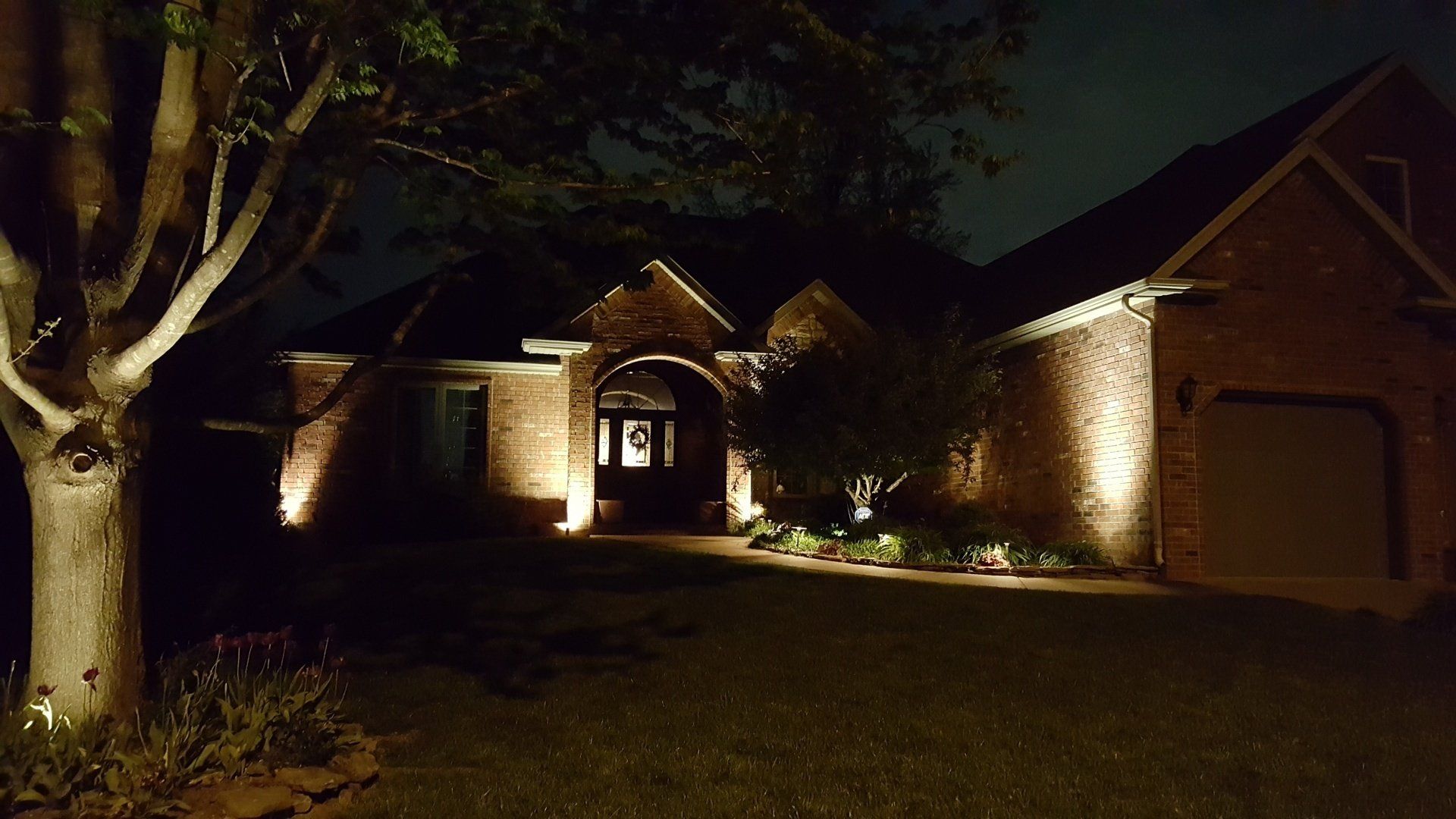 Nighttime view of a brick house with outdoor lighting illuminating the front entrance and a tree.