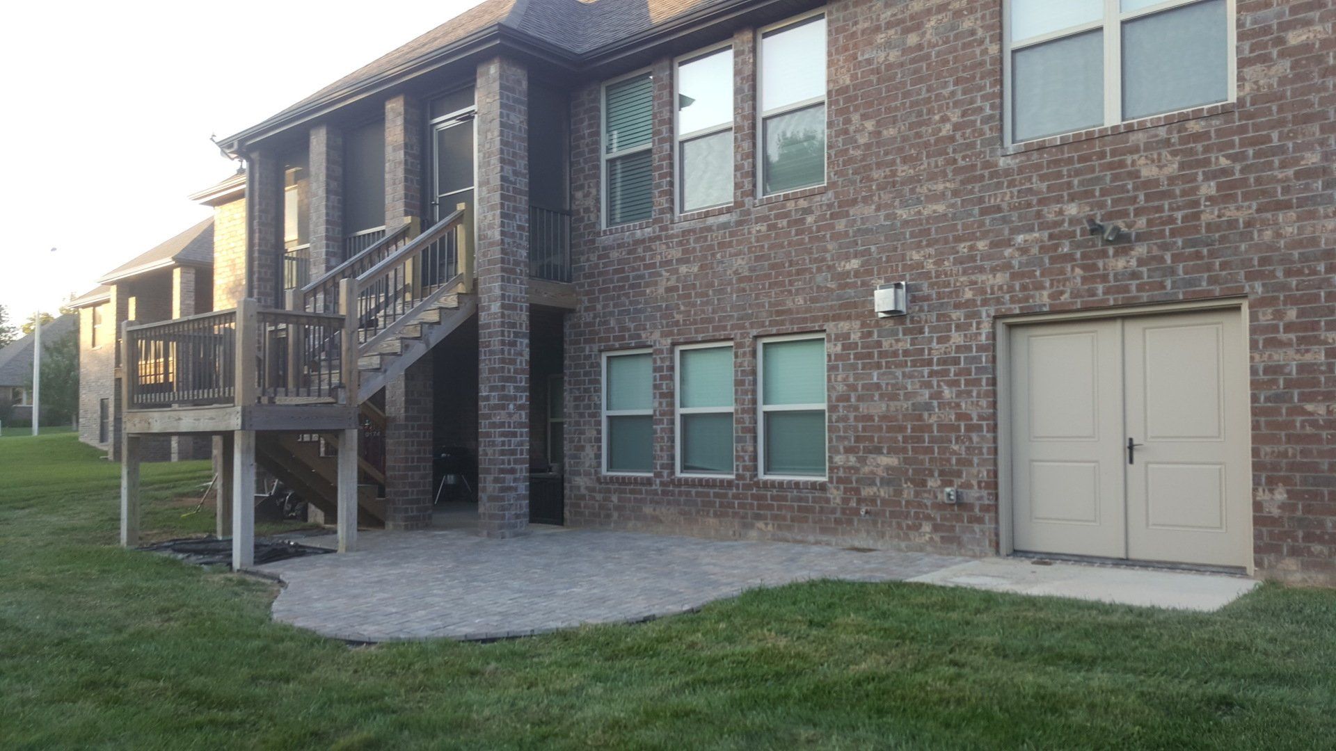 Brick house exterior with a deck, patio, and garage door. Green lawn surrounds.