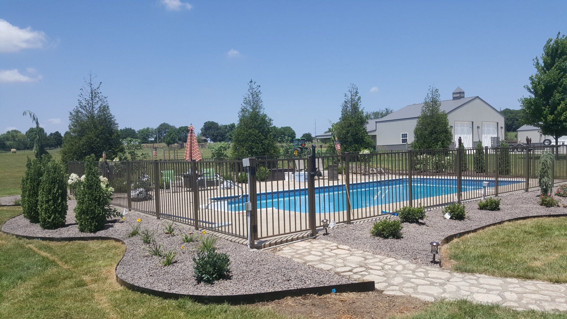 Swimming pool enclosed by a fence, surrounded by landscaping and trees on a sunny day.