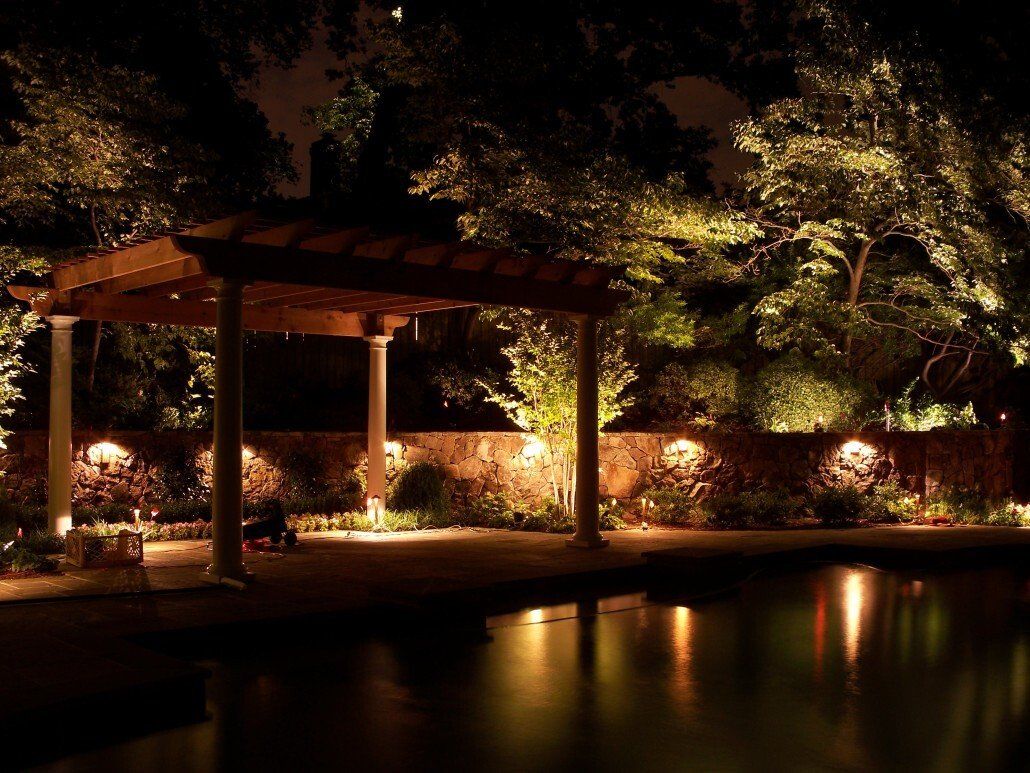 Night-lit outdoor scene with a pergola, stone wall, and a reflecting pool. Warm lights illuminate trees and structures.