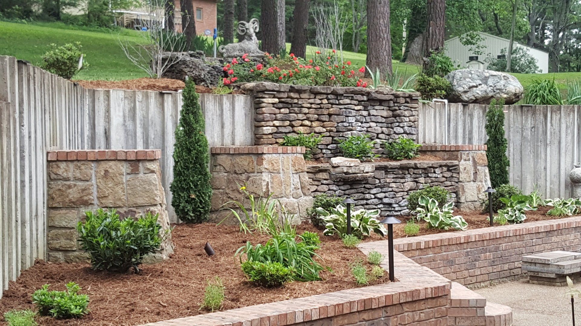 Stone tiered waterfall garden with shrubbery, bordered by a brick wall and a wooden fence.