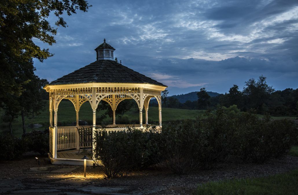 Lit gazebo at dusk, set in a garden with dark bushes and a cloudy sky.