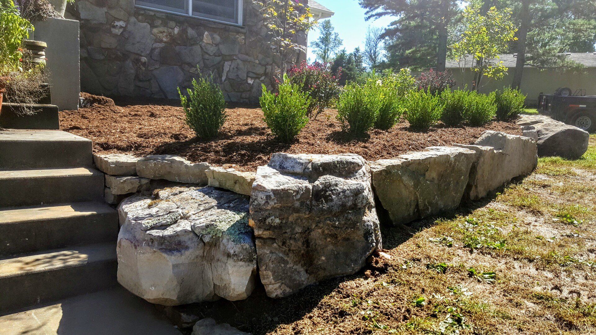 Stone retaining wall and steps leading to a house with shrubs planted in a mulched bed.