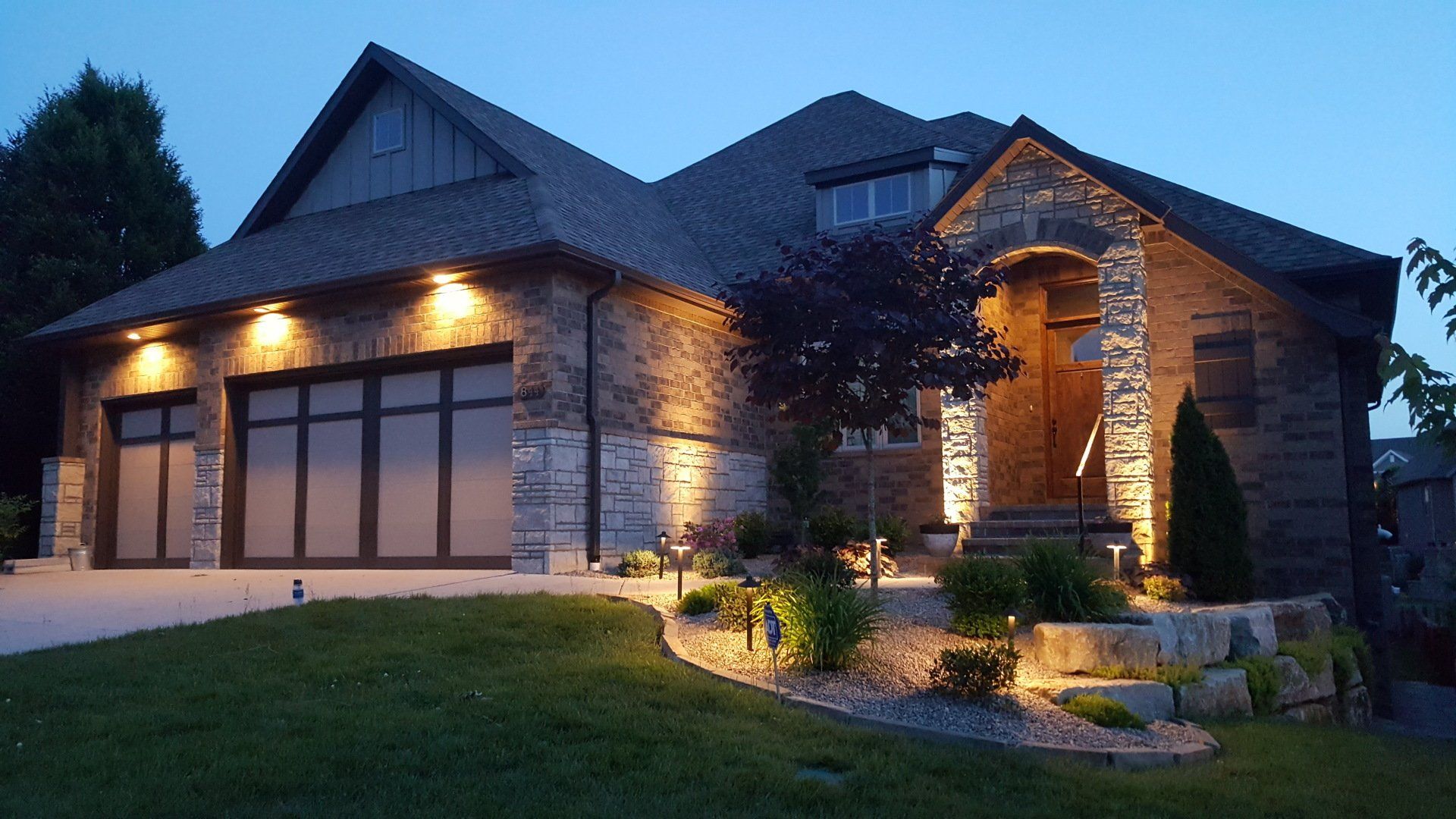 Stone house exterior lit at night, garage and entrance highlighted, landscaping with lights.