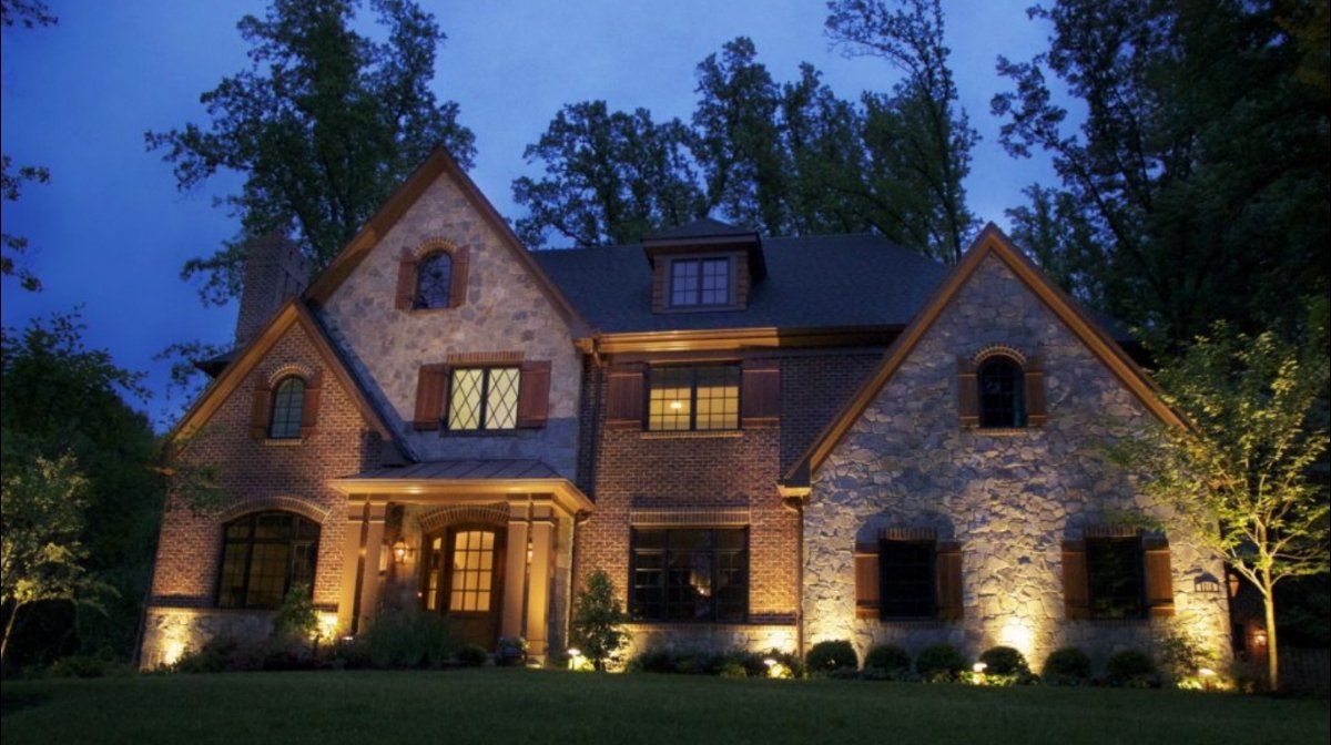 Night-lit, two-story stone and brick house with warm lighting illuminating the facade and landscaping.