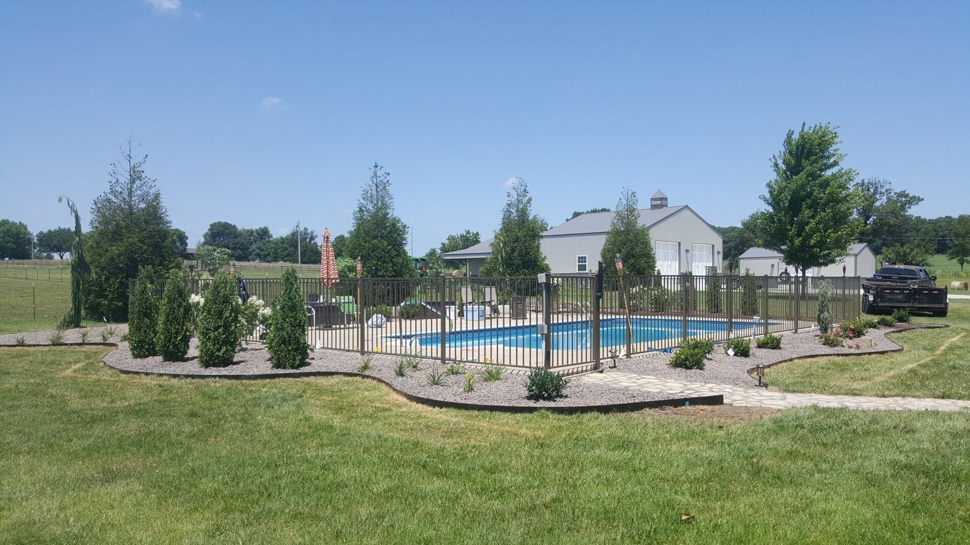 A fenced-in rectangular pool with landscaping, green grass, and a white building in the background.
