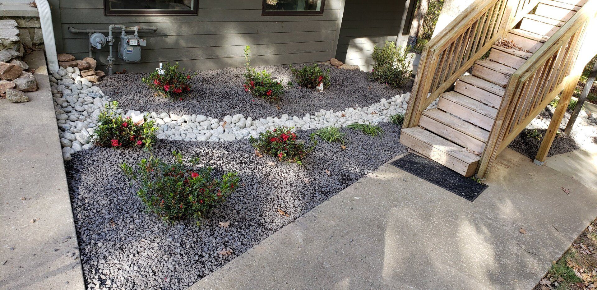 A landscaped front yard with a wooden staircase and a walkway. The garden bed has small plants and rocks.