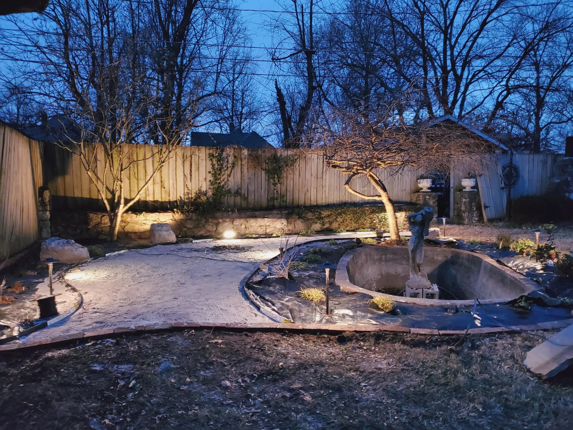 Backyard at dusk with lit pathway and pond, trees, and wooden fence.