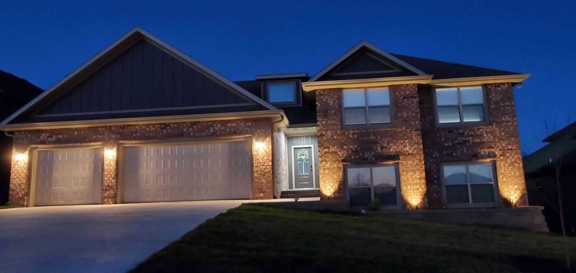 Lit-up two-story brick house at night; garage and facade spotlights illuminate exterior against dark blue sky.