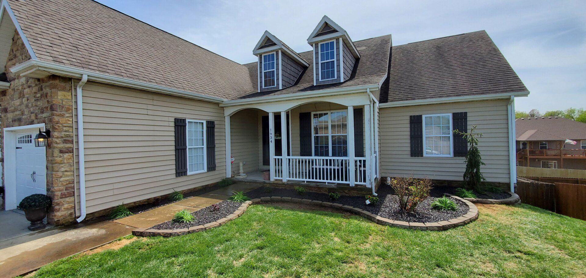 Tan house with black shutters, dormers, and a small porch. Green lawn and brick accents.