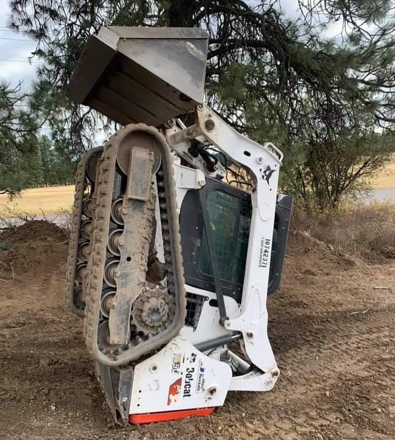 White Bobcat compact track loader with raised bucket on dirt. Tracks, trees, and sky visible.