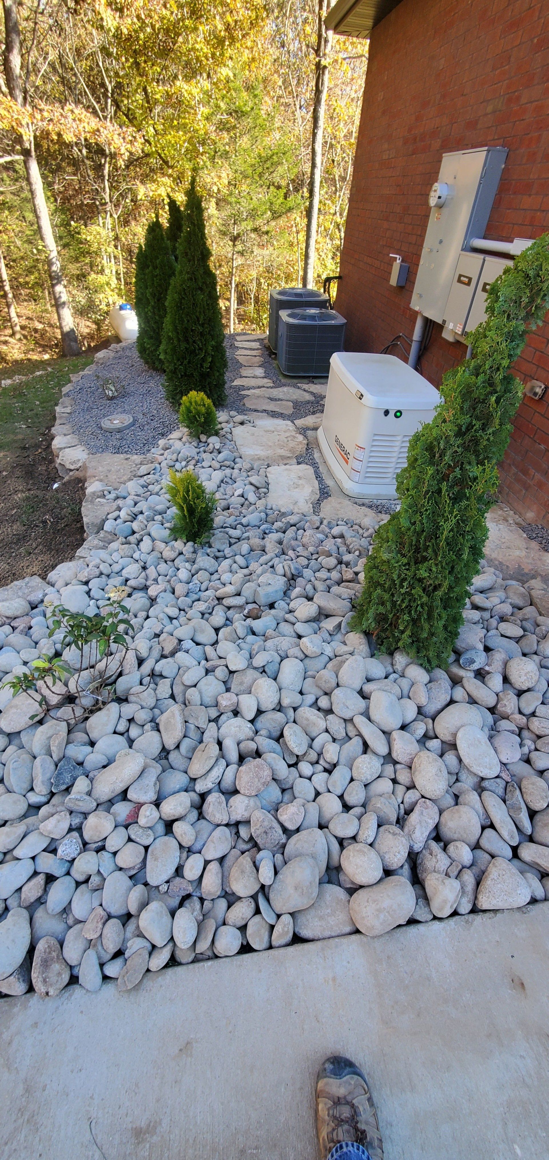 Landscaped bed with rocks, shrubs, and greenery near a brick house and utility equipment.
