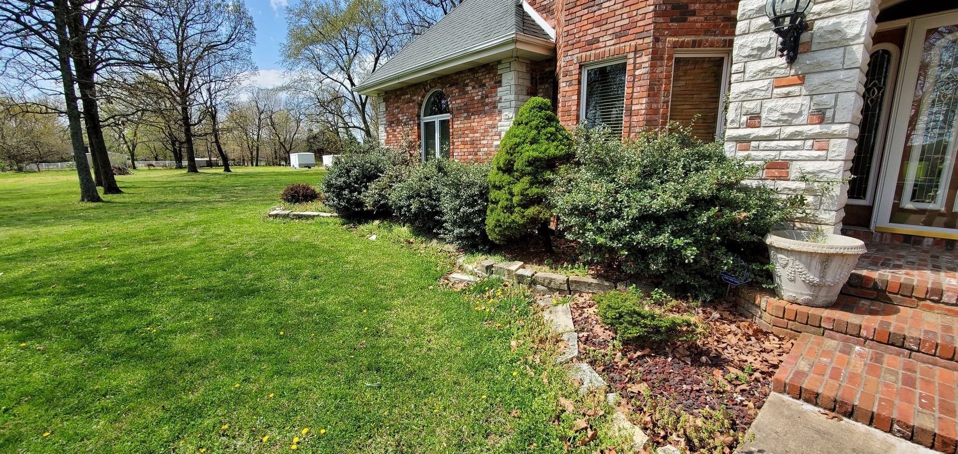 A brick house with green lawn and bushes. A stone pillar, steps, and trees are also present.