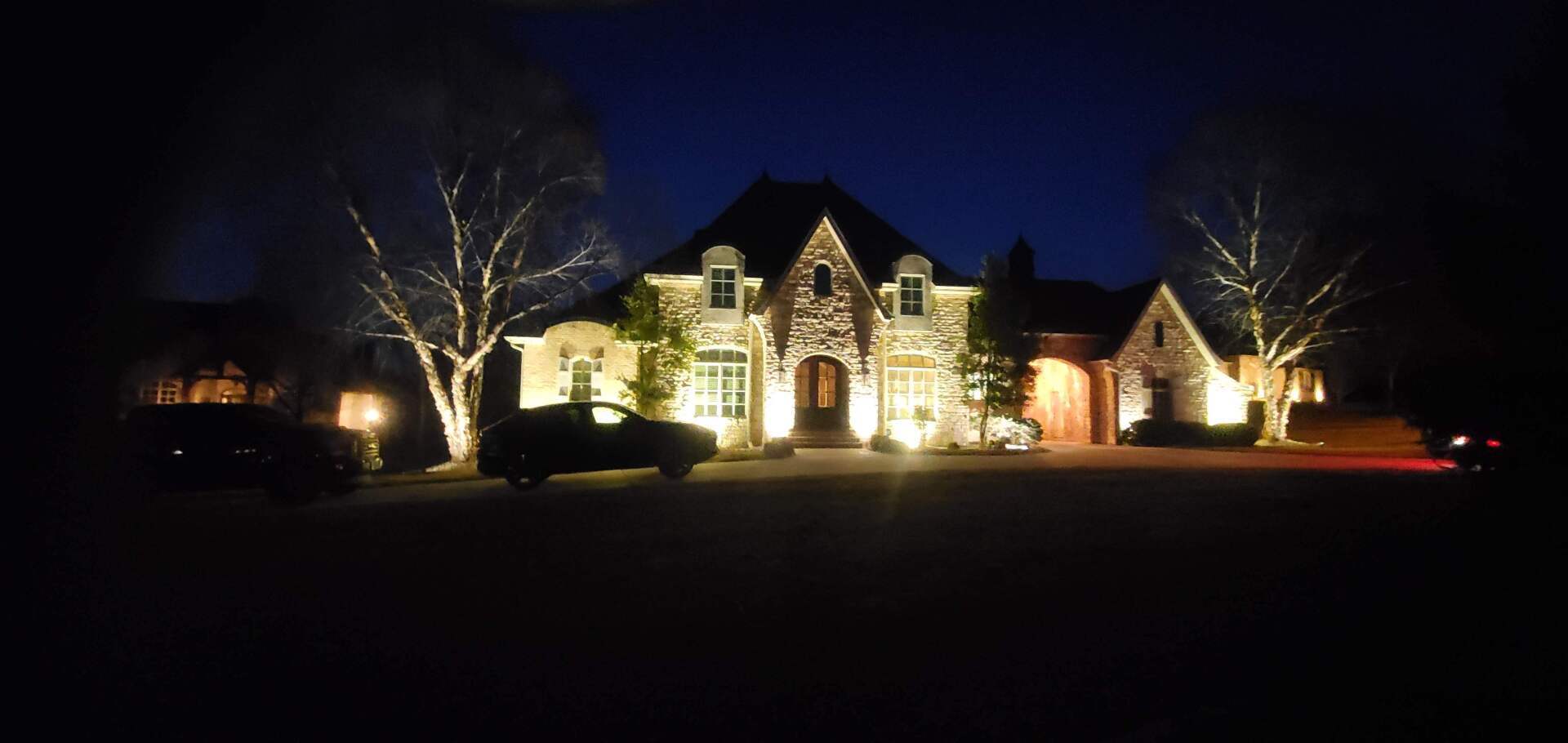 A large house lit up at night with spotlights, two cars, and trees in the foreground.
