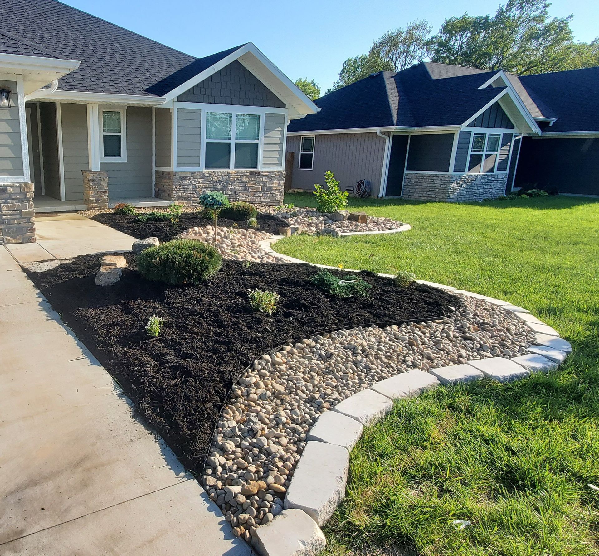 A landscaped front yard with mulch beds, stone border, and a concrete walkway leading to a house.