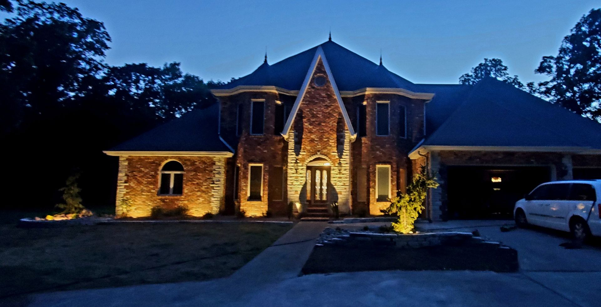 House illuminated at dusk, featuring brick facade, lit by spotlights. Driveway and trees in the background.