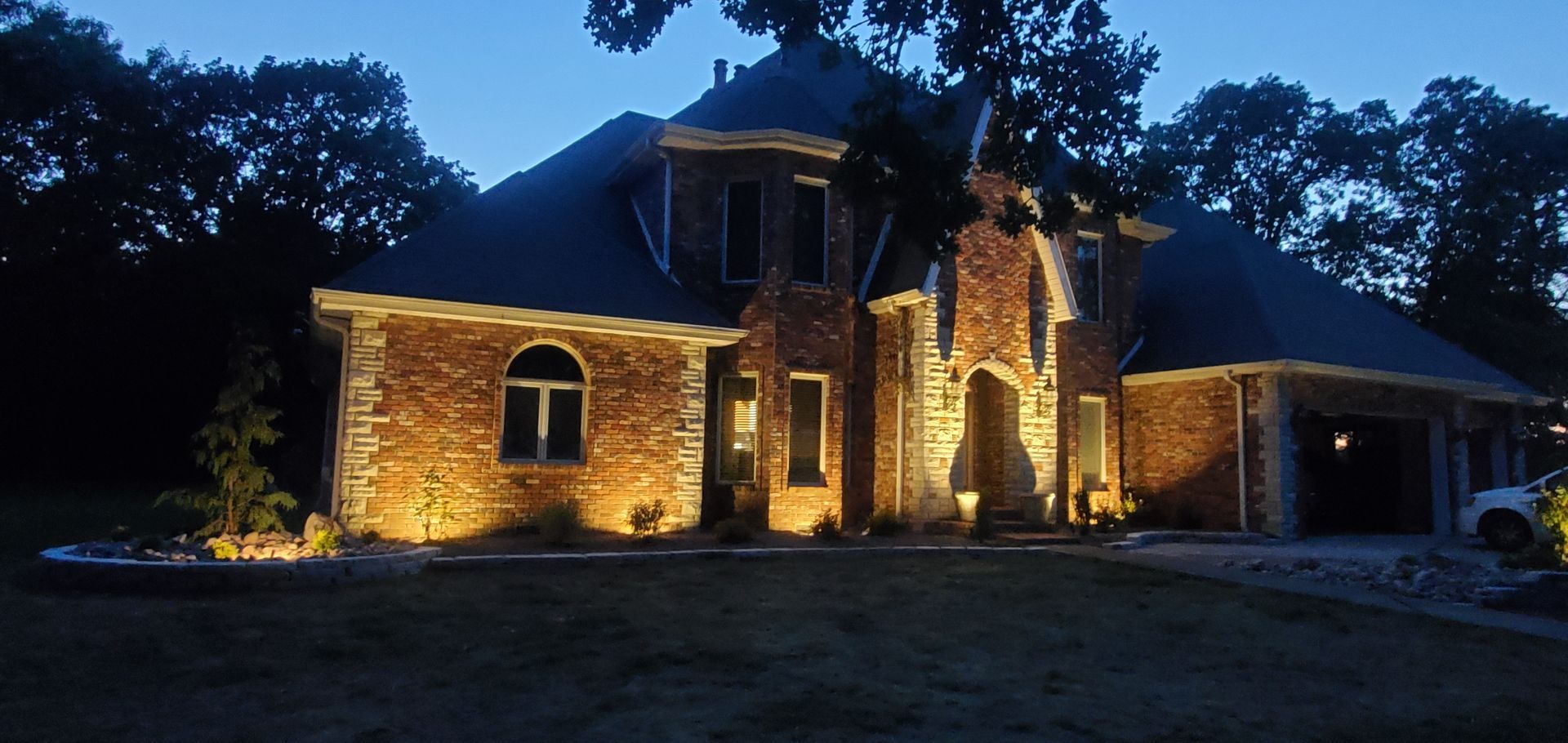 House illuminated at dusk with landscape lighting. Brick and stone facade, dark roof, trees in background.