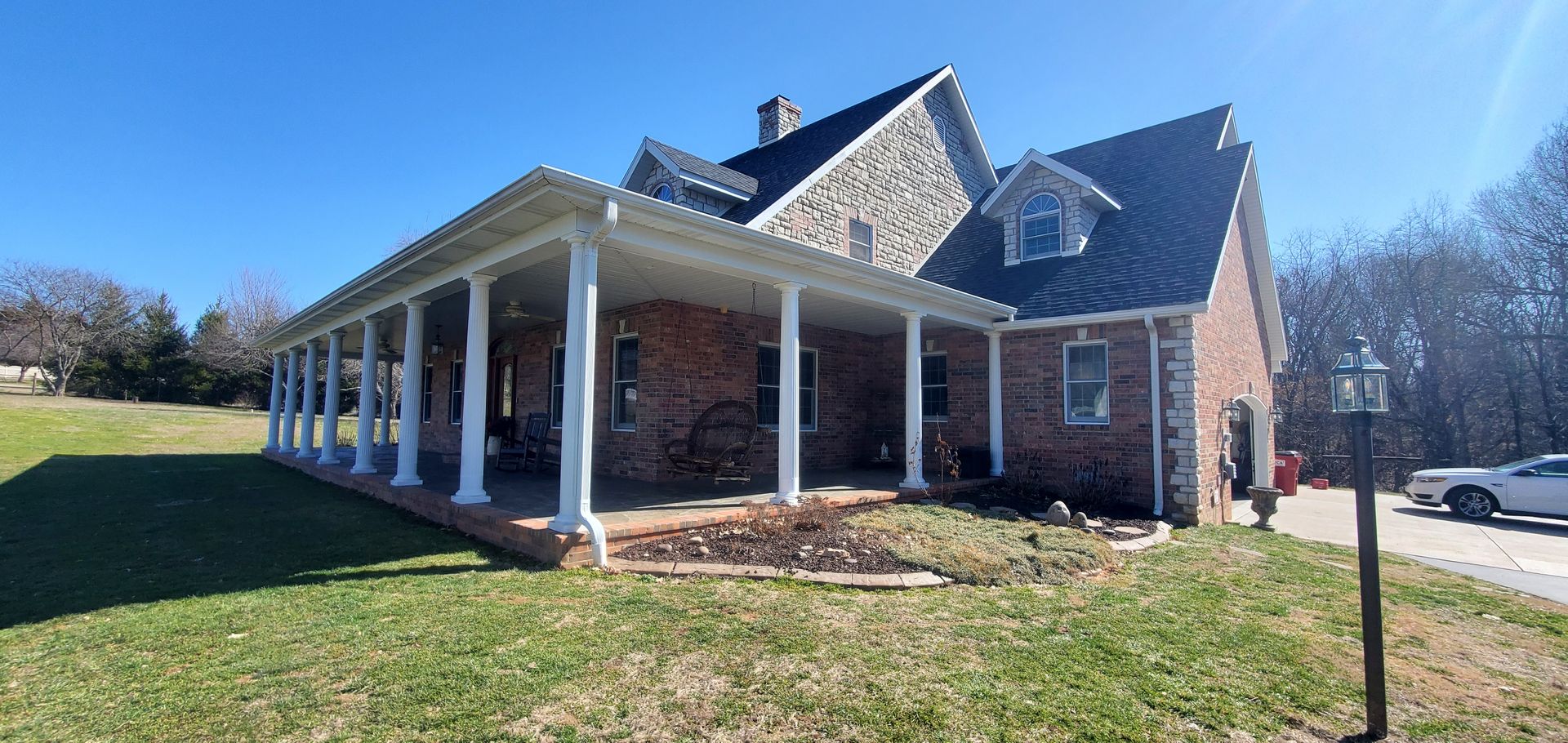 A two-story brick and stone house with a wraparound porch and columns under a clear blue sky.