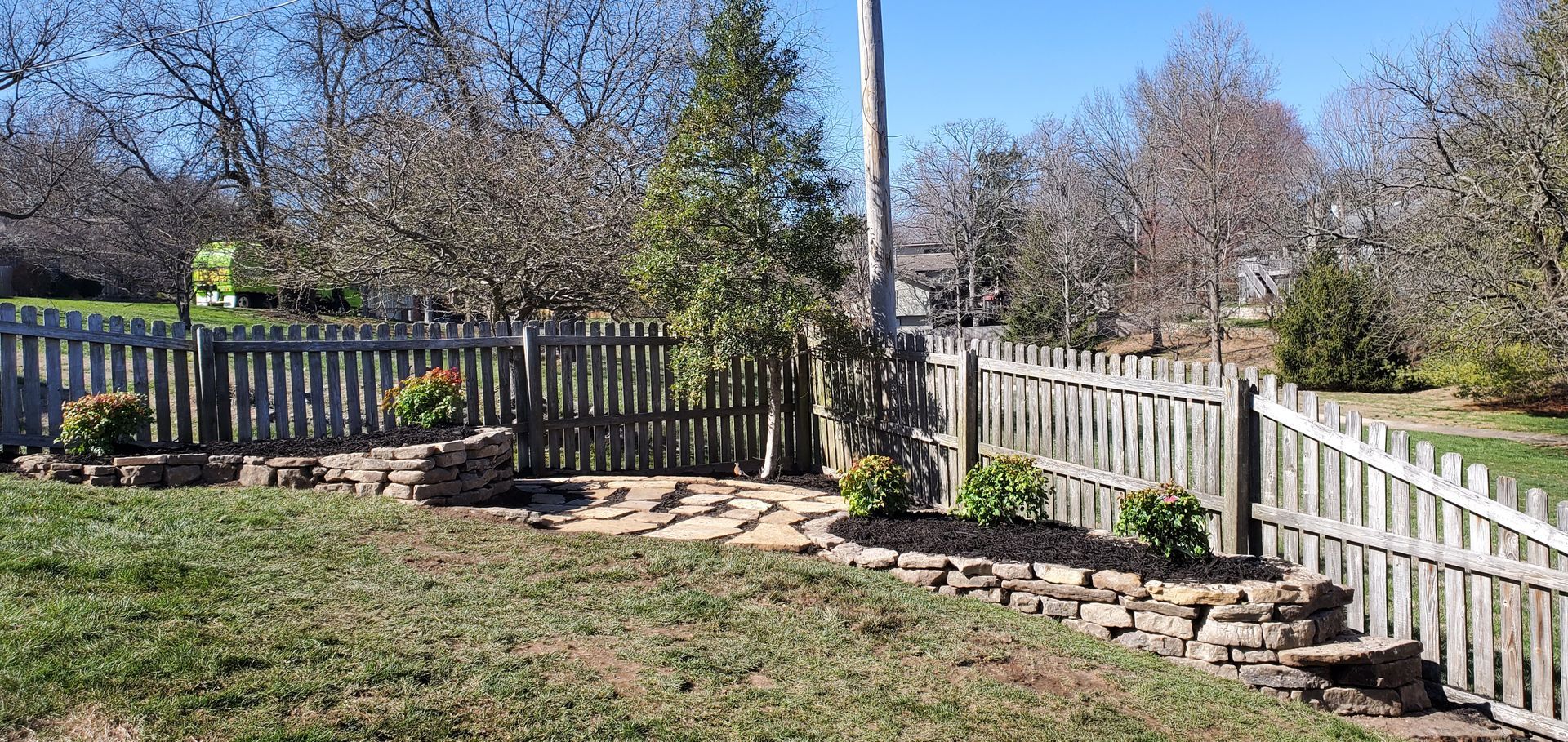 Wooden fence surrounds a yard with a stone-lined garden area and a tall pole. Trees and blue sky in the background.