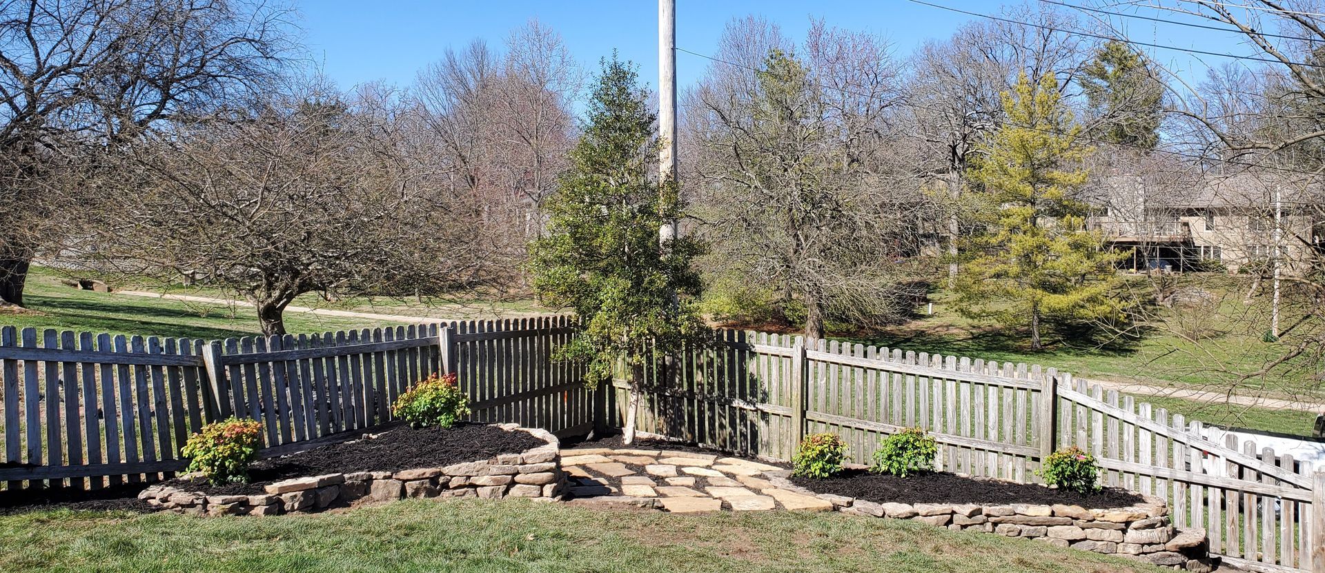A wooden fence surrounds a yard with a brick patio, green plants, and leafless trees under a blue sky.