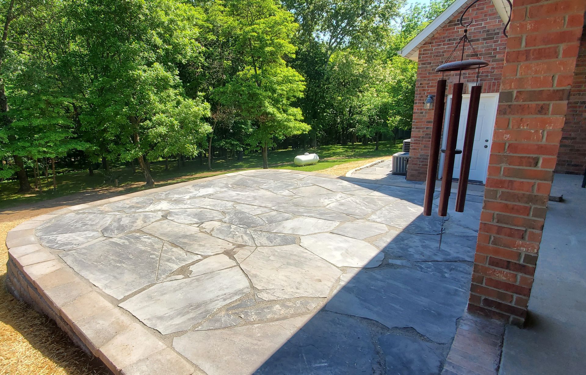 Stone patio next to a brick building and trees. Wind chimes hang on the right.