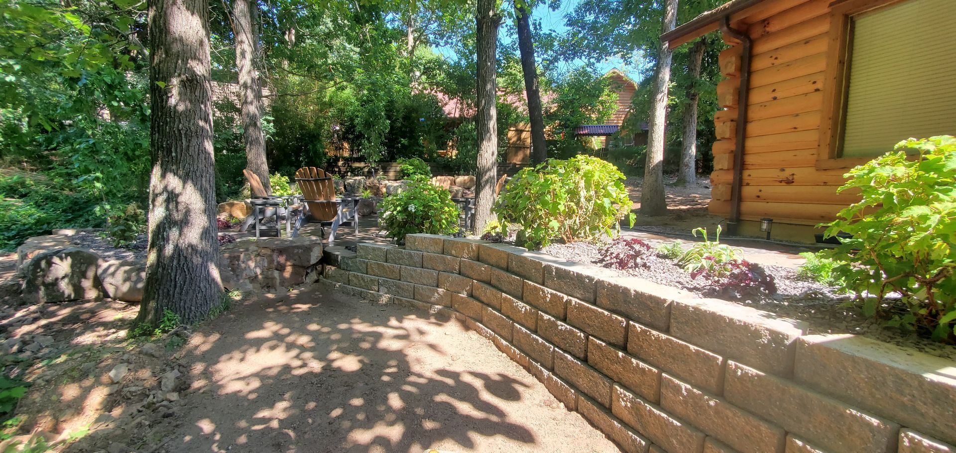 Stone retaining wall with a log cabin, trees, and lush greenery.