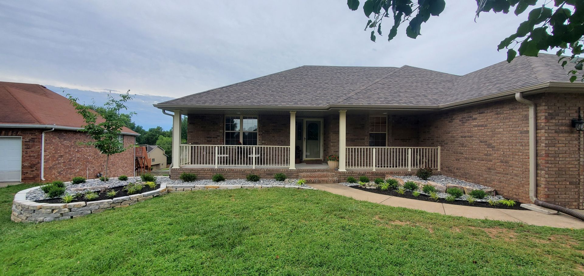 Brick house with a porch, landscaping, and a lawn on a cloudy day.