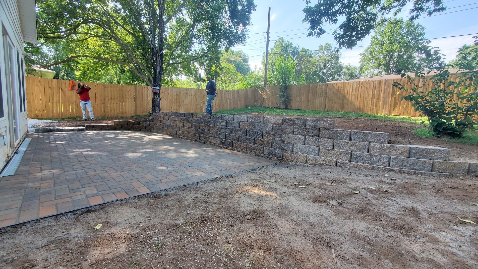 Backyard landscaping with a retaining wall, newly laid brick patio, and wooden fence. Two people are working.