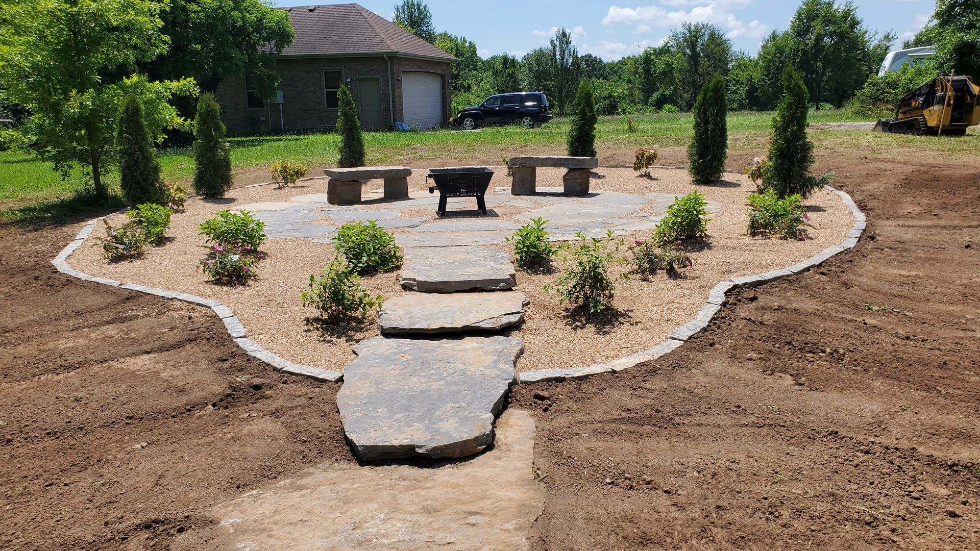Stone patio with fire pit, benches, and trees in a landscaped backyard setting.