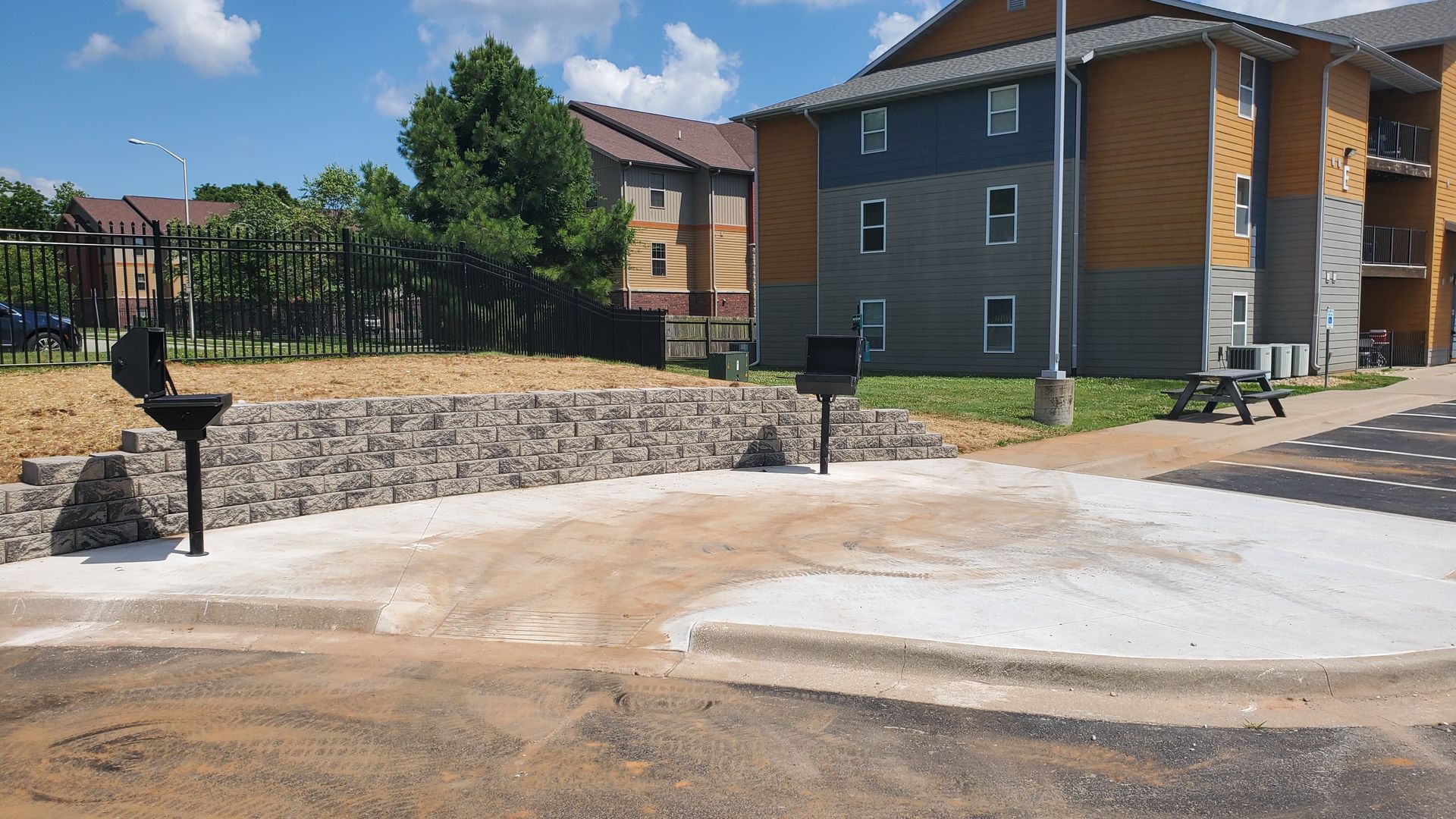 Outdoor grilling area with two grills, stone retaining wall, and apartment buildings in background.